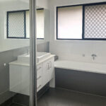 Bright contemporary bathroom with white vanity, frosted window, and built-in bathtub against dark tiled walls.
