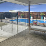 Backyard pool area with a black metal fence, blue rectangular pool, and outdoor seating with tropical cushions under a covered patio.