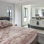 Bedroom with a pink duvet and floral pillows, a wooden bed frame on the left, and a white double-sink vanity with a large mirror on the right.