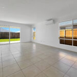 Bright empty living room with large tiled floor, white walls, and two large windows plus sliding glass doors to a fenced backyard. A wall-mounted air conditioner is visible above the right window.