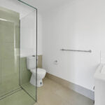 Modern bathroom with a glass-enclosed shower, toilet, and a white countertop sink on the right; neutral tiled floor and walls.