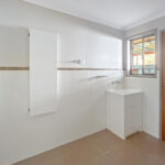 Bright laundry/bathroom with a wall cabinet, utility sink, white tiled walls, and a wooden door by a window.