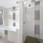 Modern bathroom with a white bathtub and a glass-enclosed shower, tiled walls and a large window on the left.