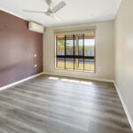 Empty living room with a purple accent wall, large window, and ceiling fan, bright natural light, light gray wood floor. (no furniture)