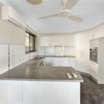 Bright white kitchen with U-shaped cabinets, gray countertop island, double sink under a window, built-in oven, and a ceiling fan.