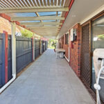 Narrow concrete walkway between brick houses under a metal canopy, with a blue wooden fence on the left and utility meters on the right.