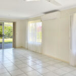 Empty living room with light tiled floor, beige walls, and a white ceiling fan; large sliding glass door with sheer curtains on the left side.