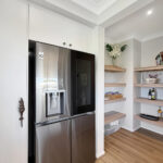 Modern kitchen area with a stainless steel French-door refrigerator flanked by white cabinets and open wooden shelves holding dishes and bottles.