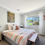 Bright bedroom with pastel striped bedding, a beige nightstand with a plant, and a large window with white curtains.