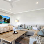 Living room with a beige sectional, wooden coffee tables, potted plants, and a TV showing a tropical beach scene.