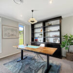 Bright home office/dining area with a black built-in shelf unit, a wooden table, and a pendant light, near large windows showing a green yard.