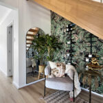 Living area under a wooden staircase with tropical wallpaper, a wall-mounted wine rack, and leafy plants beside a mirror and seating area.