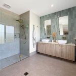 Modern bathroom with a glass-walled shower, brass fixtures, and double vessel sinks on a wooden vanity against a green-tiled wall.