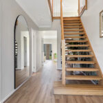 Bright entry hall with floating wooden staircase on the right and a tall curved mirror on the left.