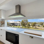 Bright modern kitchen with white cabinets, stainless range hood, and a large window overlooking a suburban street.