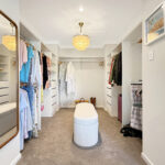 Walk-in closet with clothes hanging on both sides, white shelving, and a central white upholstered ottoman under a decorative ceiling light.