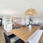 Open-concept living area with a wooden dining table, woven pendant light, white kitchen and a gray sofa.