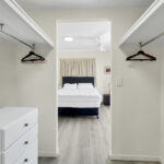 Walk-in closet opening to a bedroom with a white dresser on the left, and rods with hangers along both walls leading to a bed in the adjacent room.