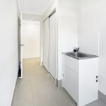 Narrow hallway with white walls and a white cabinet with stainless sink on the right side, leading to another room with an open door.
