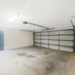 Empty residential garage with a large metal sectional door, concrete floor, and sliding glass door to the outside patio area.