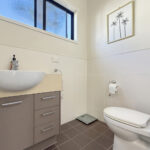 Modern bathroom with a beige vanity, round white sink, gray drawers, wall scale, and a toilet; palm-tree artwork on the tiled wall.