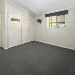 An empty room with white built-in closet doors, a wall-mounted oscillating fan, and a window with a security screen on beige walls.