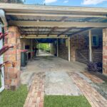 Covered carport with brick columns, metal beams, and a concrete brick-paver path bordered by tropical plants on both sides.