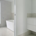 Modern white bathroom with a freestanding tub beside a frosted window and glass shower door, in a minimal space.