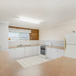 Bright kitchen with white cabinets, beige countertops, and a double sink under a window on a brick accent wall.