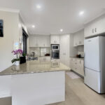 Modern white kitchen with an island, marble countertop, orchid plant, white cabinets, and stainless steel fridge with double ovens.