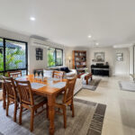 Bright open-plan living and dining room with a wooden dining table, beige sofa, bookshelf, and piano against a light wall. Large windows with screens offer garden views.
