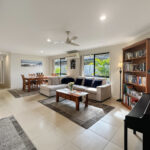 Bright open-plan living room with beige walls, white tiled floor, a beige sectional sofa, coffee table, piano, and tall bookshelf.