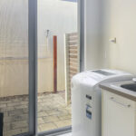 Laundry room with top-loading washing machine next to a stainless sink and countertop. Sliding glass door opens to a small patio with a fence and wooden post outside.