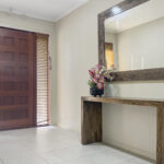 Entryway with a wooden front door, blinds, and a white tiled floor; a rustic console holds a pink flower arrangement beside a large square mirror.