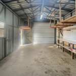 A metal-walled storage shed with long workbenches on the right and a rolling shutter at the far end, illuminated by a ceiling light.