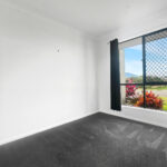Bright empty bedroom with dark carpet and a large window with a security screen and black curtains, showing a street and plants outside.