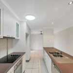 Long white kitchen with brown countertops, upper cabinets, stovetop, and a double sink along the right wall.