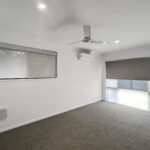 Empty living room with white walls, ceiling fan, wall-mounted AC, and large glass door with roller blinds on a gray carpeted floor, minimal decor