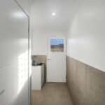 Narrow laundry/utility room with a sink at the far end, door with a small barred window, and beige-tiled lower walls.