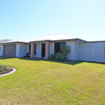 Front view of a single-story suburban house with a two-car garage, white exterior, and a well-kept green lawn with desert plants.