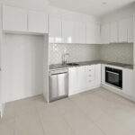 Modern white kitchen with built‑in cabinets, stainless dishwasher, and stainless oven along an L-shaped counter with a herringbone tile backsplash