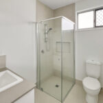 Modern bathroom with a glass shower enclosure, white toilet, and rectangular sink on a beige countertop near a frosted window with a security grill.